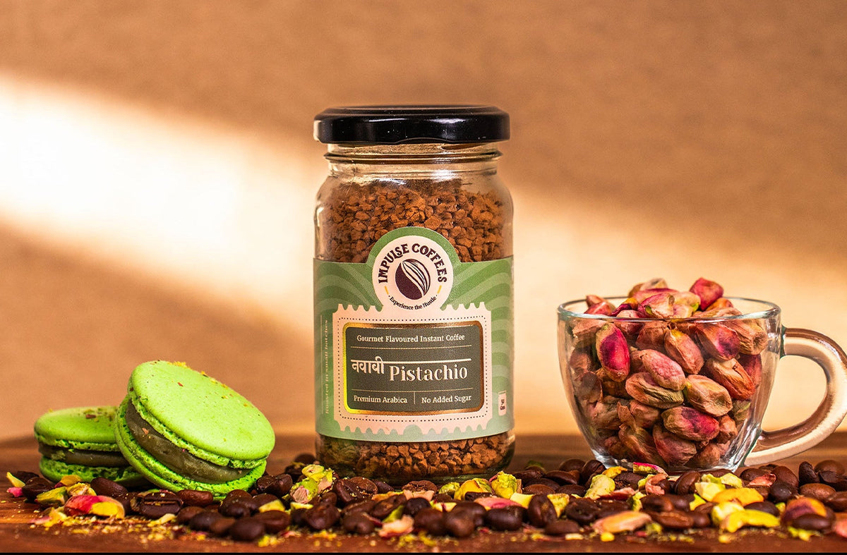 A jar of pistachio-flavored instant coffee labeled “Impulse Coffees” is displayed on a wooden surface, surrounded by coffee beans, green pistachio macarons, and a glass cup filled with whole pistachios, with a warm, soft-focus background.